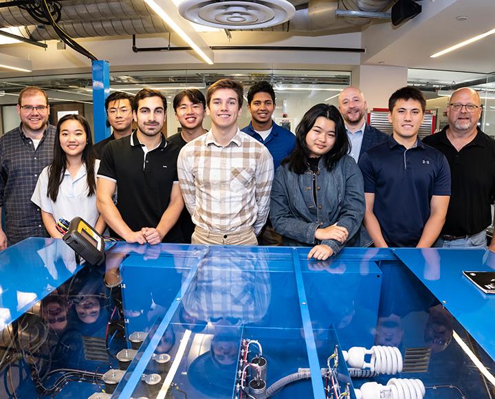 A group of students stand behind a clear-top blue table that contains electrical equipment