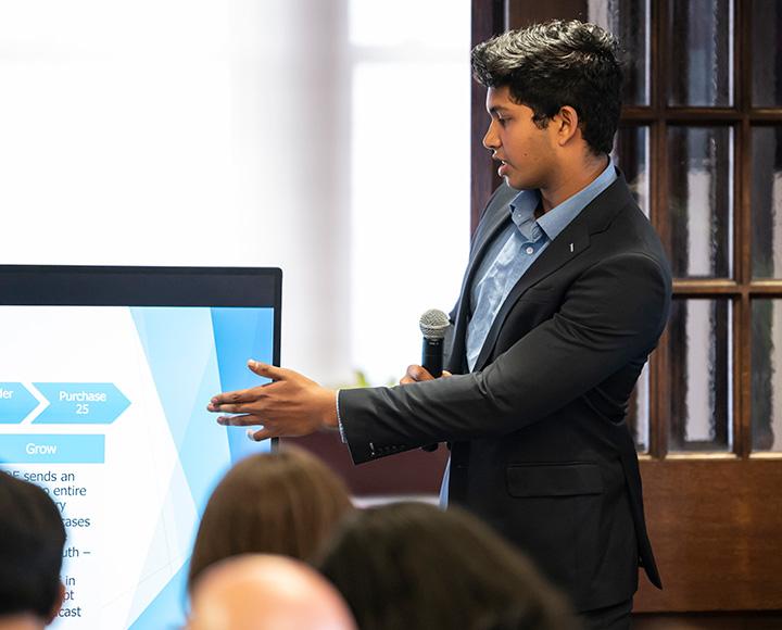A person in a suit points to a screen as he gives a presentation