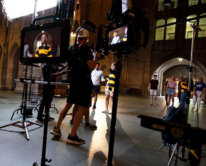Monitors on tripods show a person in a striped polo. The film set is in the background.