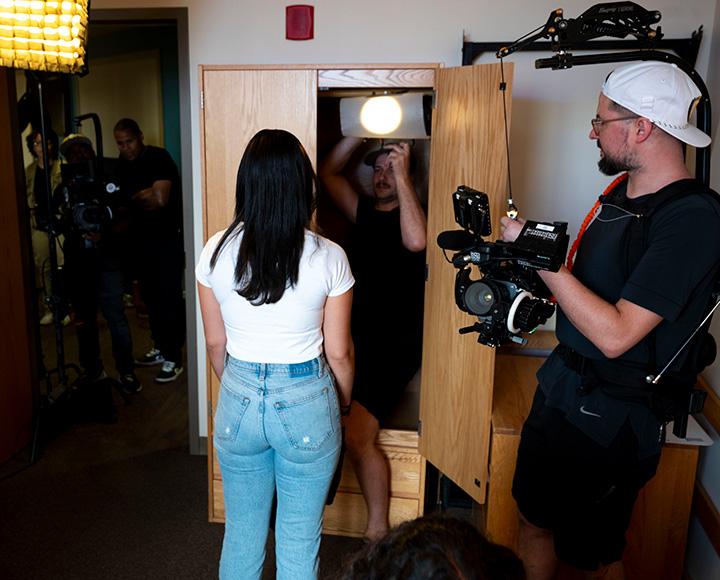 People film a person standing in a dorm room. A lighting operator stands in an open wardrobe.