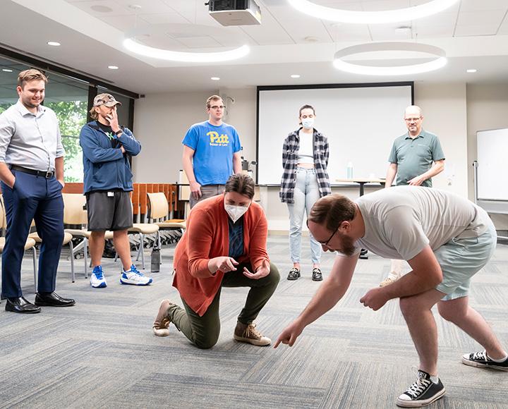 Two students kneel while performing a scene. Other students stand in a circle around them.