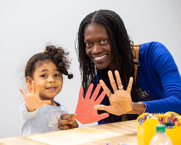 A young girl and a woman hold up their hands. The woman is also holding a construction paper hand cutout.