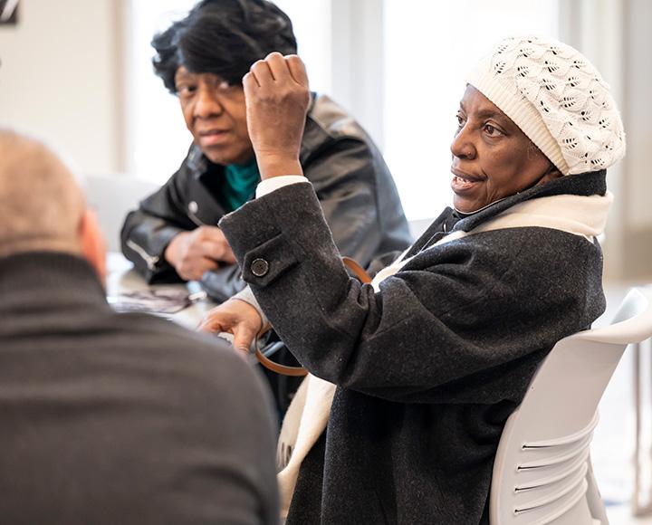 A person, holding up one hand, speaks to others sitting around a table