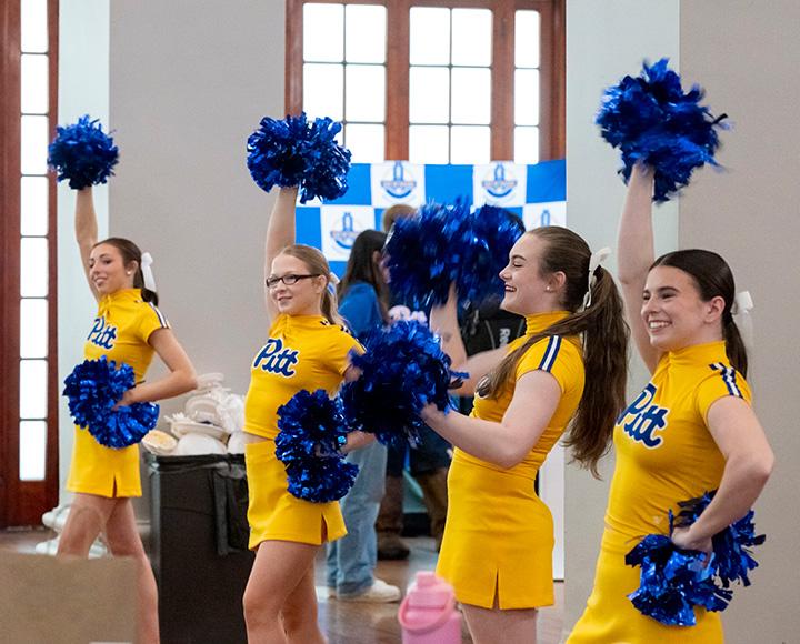 Four cheerleaders in yellow Pitt uniforms wave blue pompoms