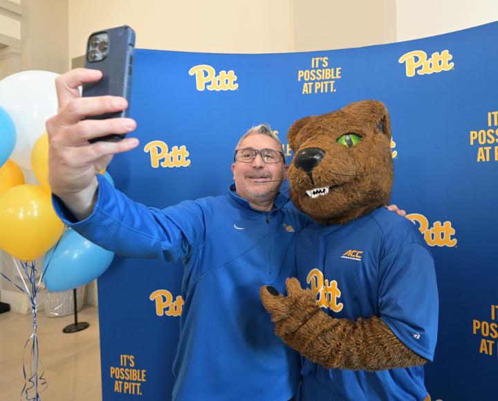 Evan Facher takes a selfie with Roc the Panther in front of a Pitt-branded background