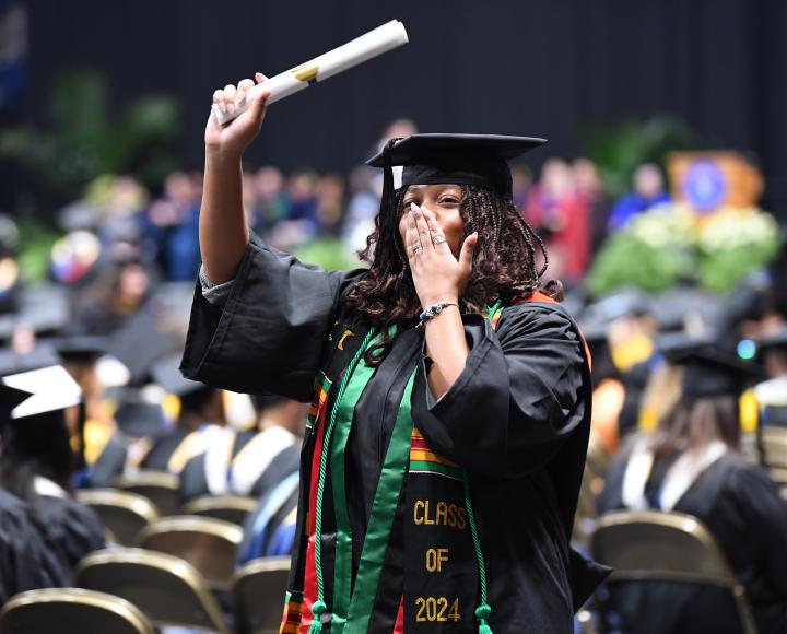 A graduate holds her diploma in the air while blowing a kiss to audience members.