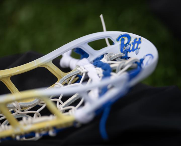 Closeup of a gold and white lacrosse stick head adorned with the blue and gold script Pitt logo