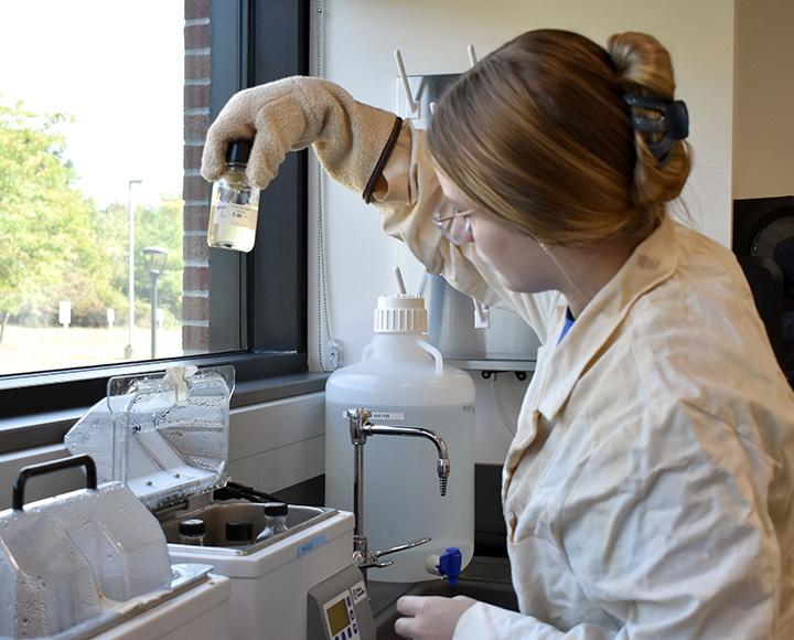 A student in a lab coat holds up a bottle of clear liquid to a window's light.