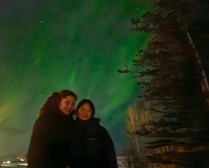 Two people stand in front of the Northern Lights.