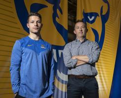 A person in a blue shirt and a person in a checkered shirt stand in front of a yellow and blue panther banner