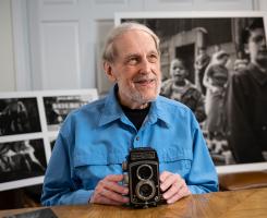 Rosella sits with his camera in front of photographic prints