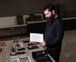 Ben Rubin, Horror Studies Collection Coordinator at the University Library System, stands in a darkly lit room before a table of newly acquired Blair With Project items in the Special Collections section of the Hillman Library. Items include film posters, props, pamphlets and more.