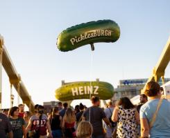 Many people on one of the Pittsburgh bridges, a child carries a pickle-shaped balloon that reads "PicklesBURGH".  In the distance there is a pickle-shaped sign reading "HEINZ".