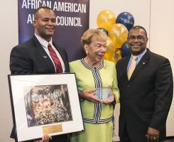 A woman receives an award while standing next two two men in front of a sign that reads "African American Alumni Council"