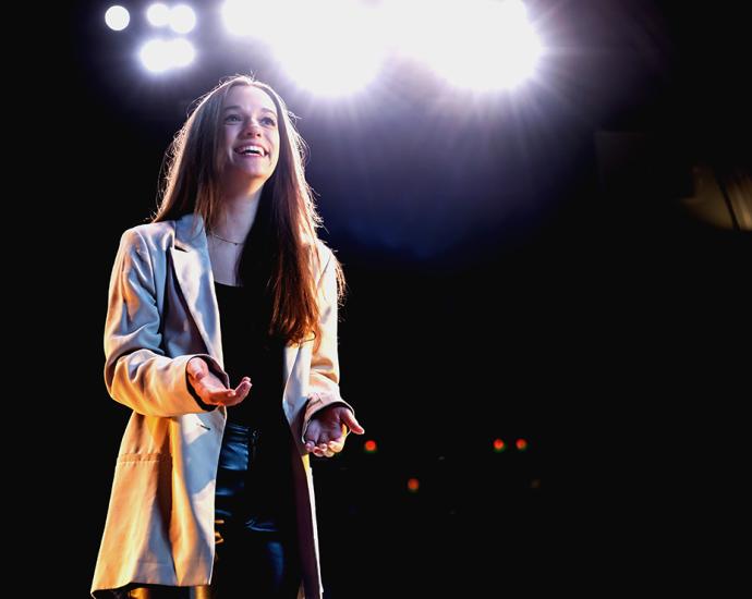 A woman smiles while standing on a stage in an empty theater as spotlights shine behind her