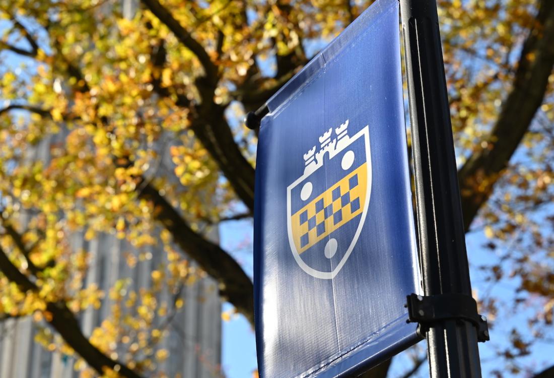 Banner with Pitt shield flies in front of fall leaves and Cathedral of Learning.