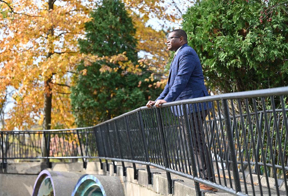 Means leans on a railing in a park, surrounded by fall leaves