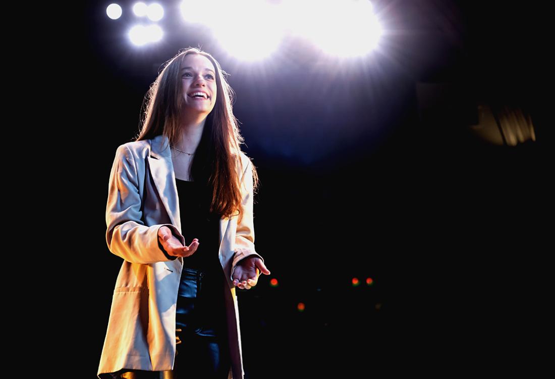 A woman smiles while standing on a stage in an empty theater as spotlights shine behind her