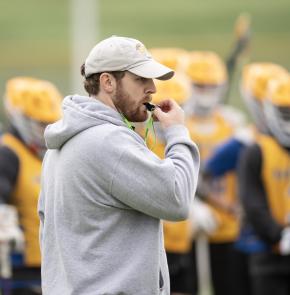 A coach stands in front of his lacrosse team and blows a whistle