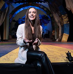 ​​A woman gestures with her hands while smiling as she sits on the end of a stage in an otherwise empty theater