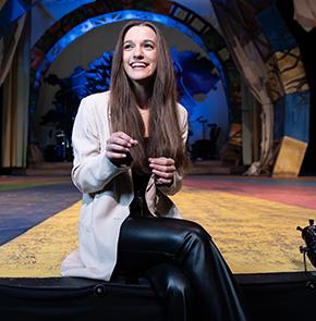​​A woman gestures with her hands while smiling as she sits on the end of a stage in an otherwise empty theater