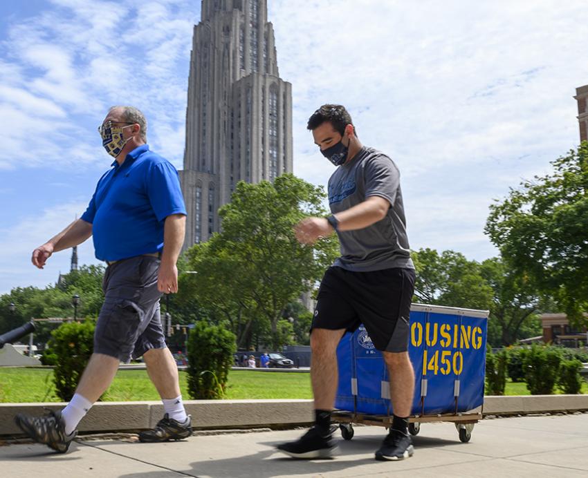 Two men in masks walking with a housing cart with the Cathedral of Learning in the background