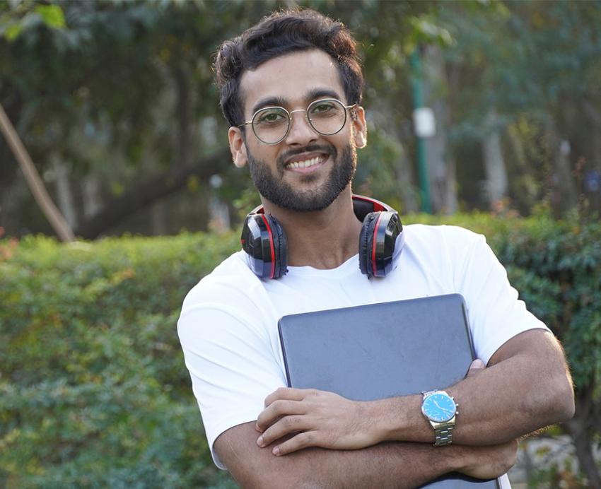 Student in white shirt holding laptop in front of shrubbery