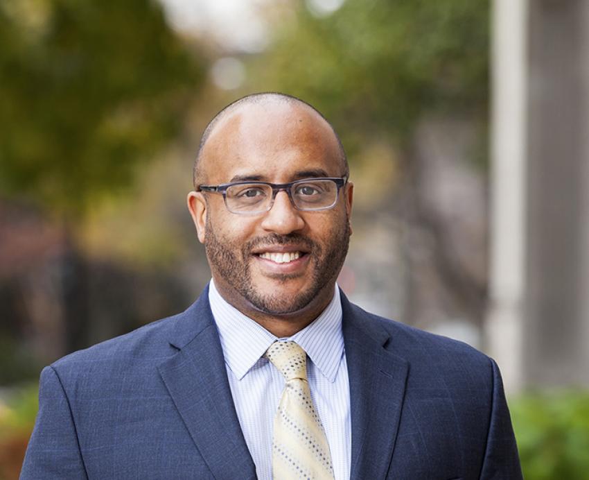 James Huguley smiling with glasses, navy blue suit and gold tie