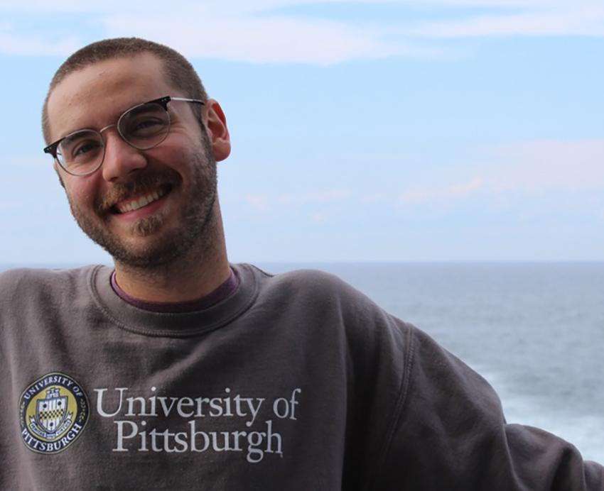Brett Bankson smiling with grey Pitt sweater, ocean in the background