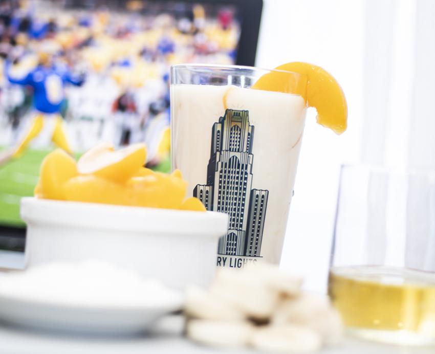 Peach smoothie with bowl of sliced peaches in front of laptop showing photo of Pitt football game