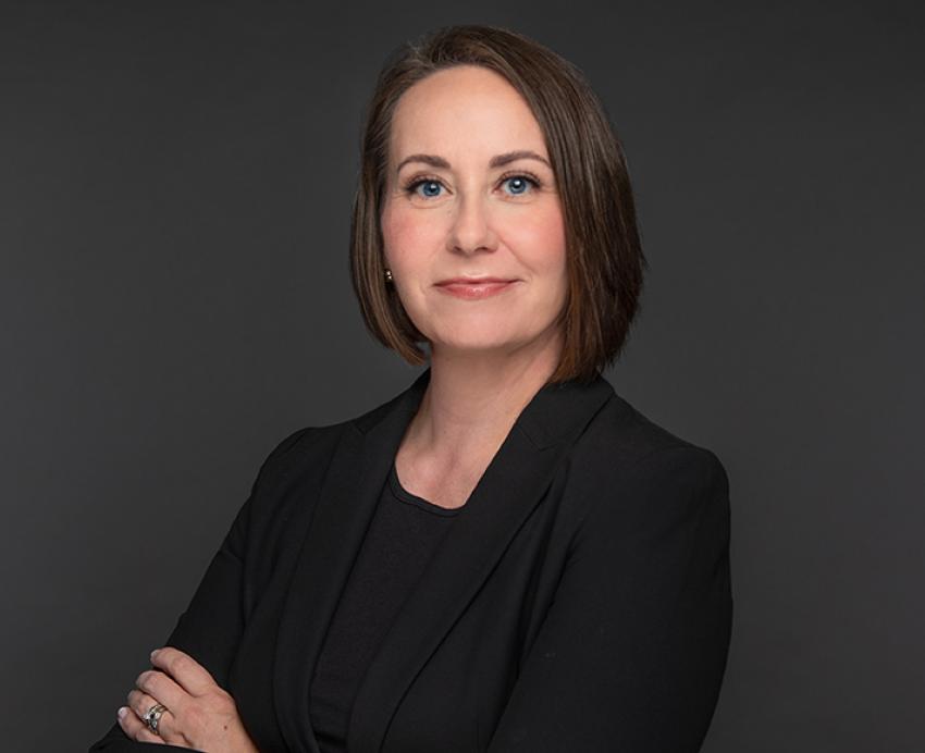 Lina Dostilio in black suit with arms folded in front of grey background