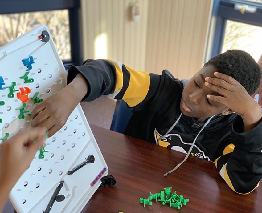 A child in a black and gold sweatshirt reaches for a board on a table