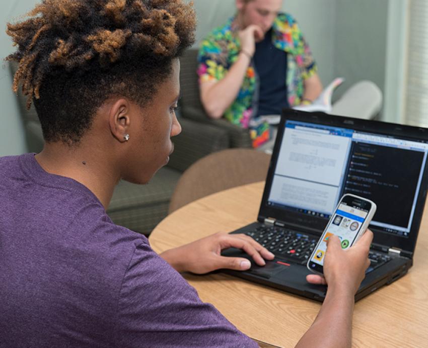 a student at a laptop holding a phone