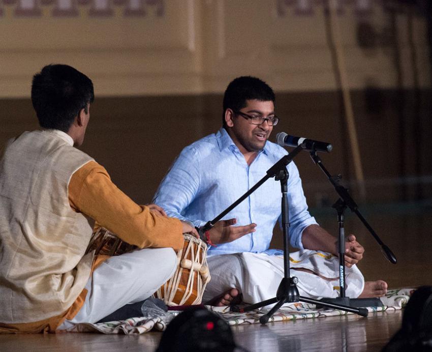 One person drums and another sings while sitting on the floor in front of microphones
