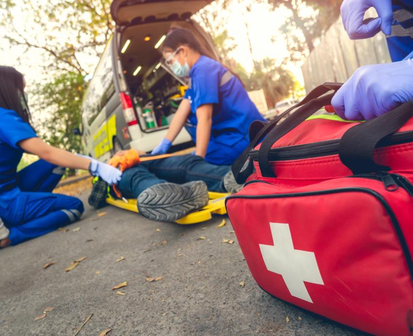 A gloved hand holds a red bag with a white cross as two people in scrubs help a person on the ground.