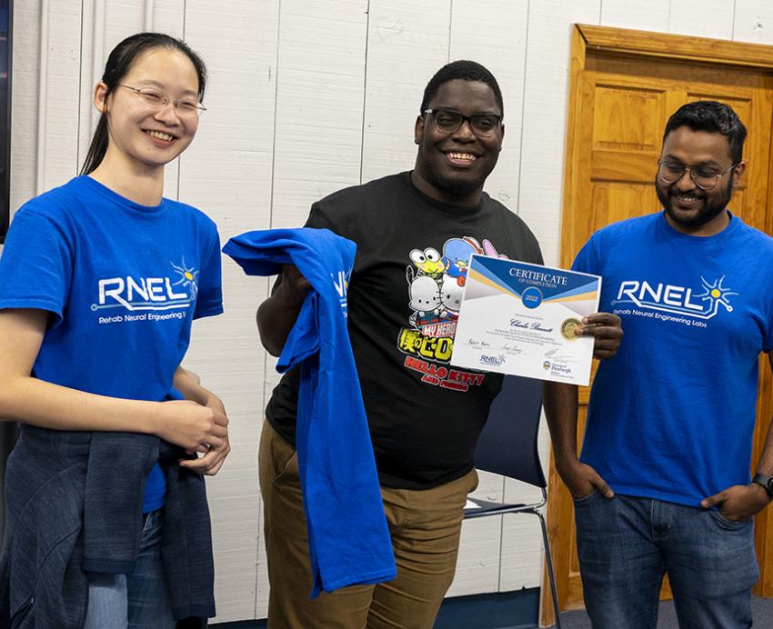 A person holds a certificate and T-shirt while standing in between two people in the same blue T-shirt that reads RNEL