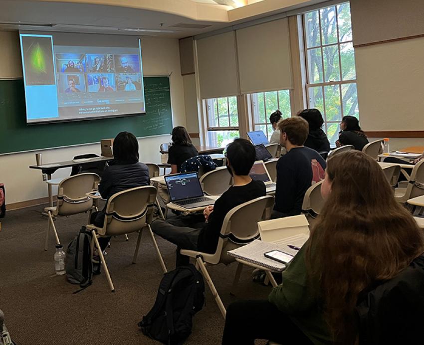 Students in rows of desks watch a video call on a projector