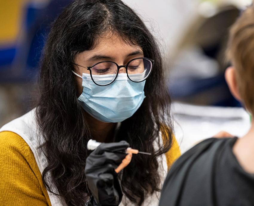 A person in glasses and a blue face mask holds a syringe in a gloved hand