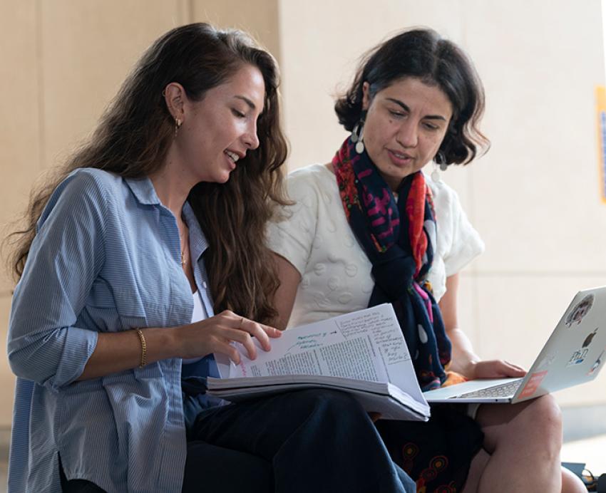 A person in a blue shirt shows a paper to someone in a white shirt and scarf holding a laptop