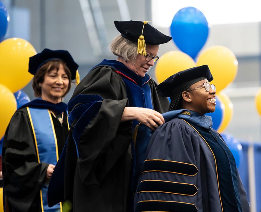 A person in doctoral regalia is hooded in front of blue and yellow balloons
