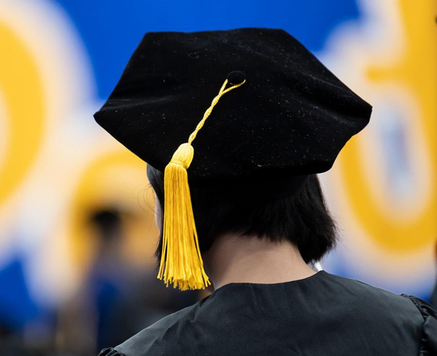 A person stands facing away, with a black velvet tam with a yellow cord on their head