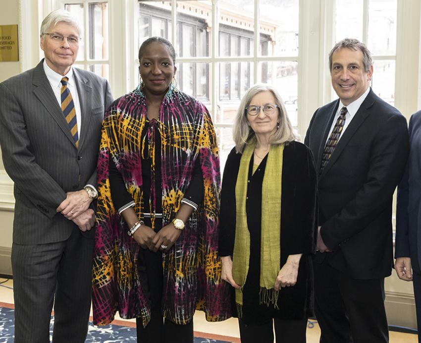 Three people in suits stand with a person in a colorful shawl and a person in a black shirt and green scarf.