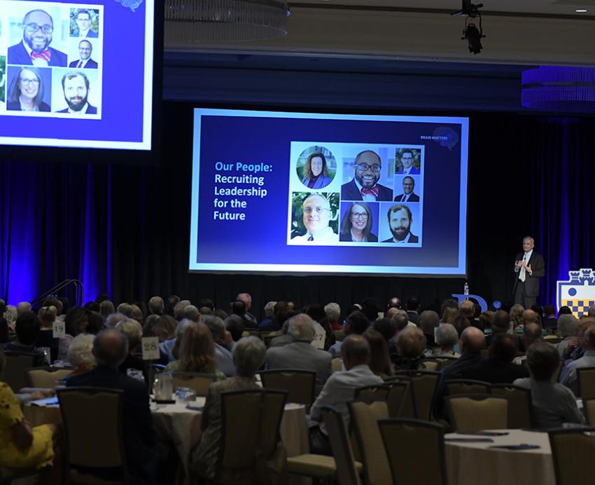 People sit at tables in a ballroom watching a presentation