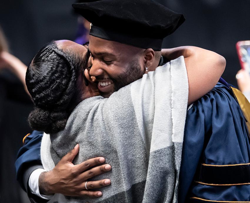 A person in blue doctoral regalia hugs someone in a grey shawl