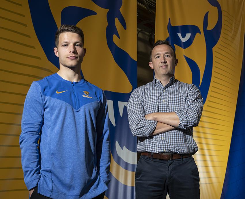 A person in a blue shirt and a person in a checkered shirt stand in front of a yellow and blue panther banner