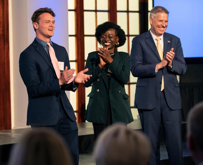 A person in a tan jacket laughs next to three people clapping in suits