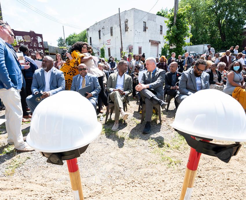 A crowd sits in chairs in front of a row of ceremonial shovels and hardhats