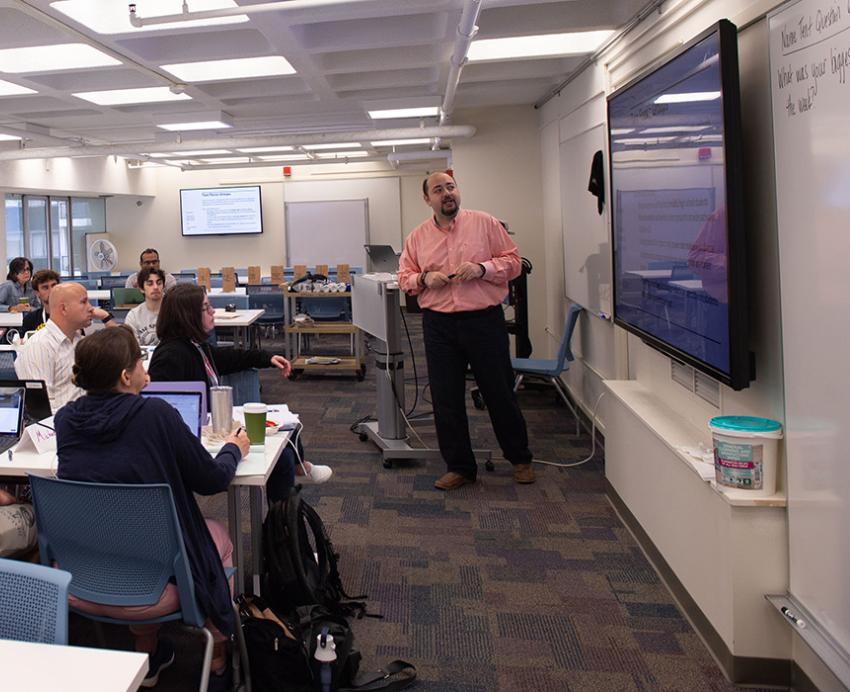 People sit with laptops in a classroom, watching a person present on a large screen