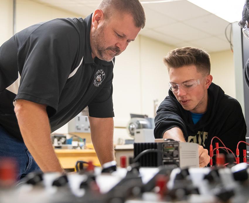 A professor talks to two students working on electrical equipment