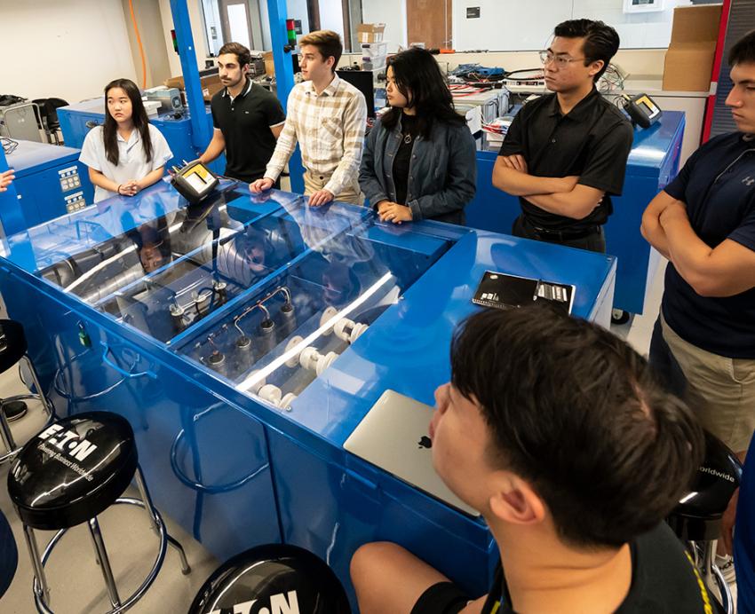 Students gather around a blue clear-top table in a lab and listen to an instructor